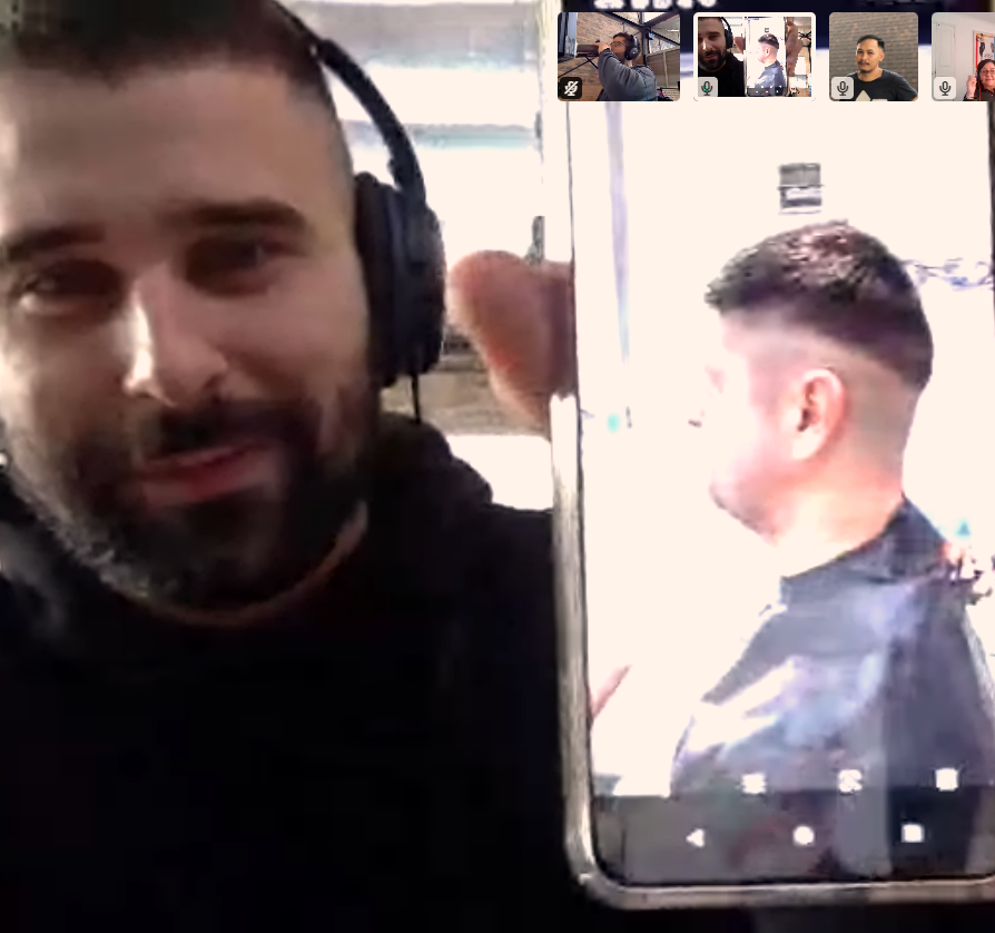 A man wearing headphones holds a smartphone showing a side view of his freshly cut short hairstyle during a video call with four other participants visible in small windows at the top of the screen.