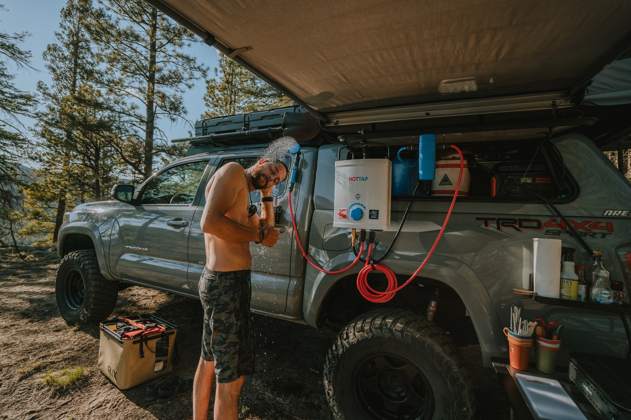 Man using an outdoor portable hot water shower system mounted on a rugged off-road vehicle at a wooded campsite with camping supplies nearby.