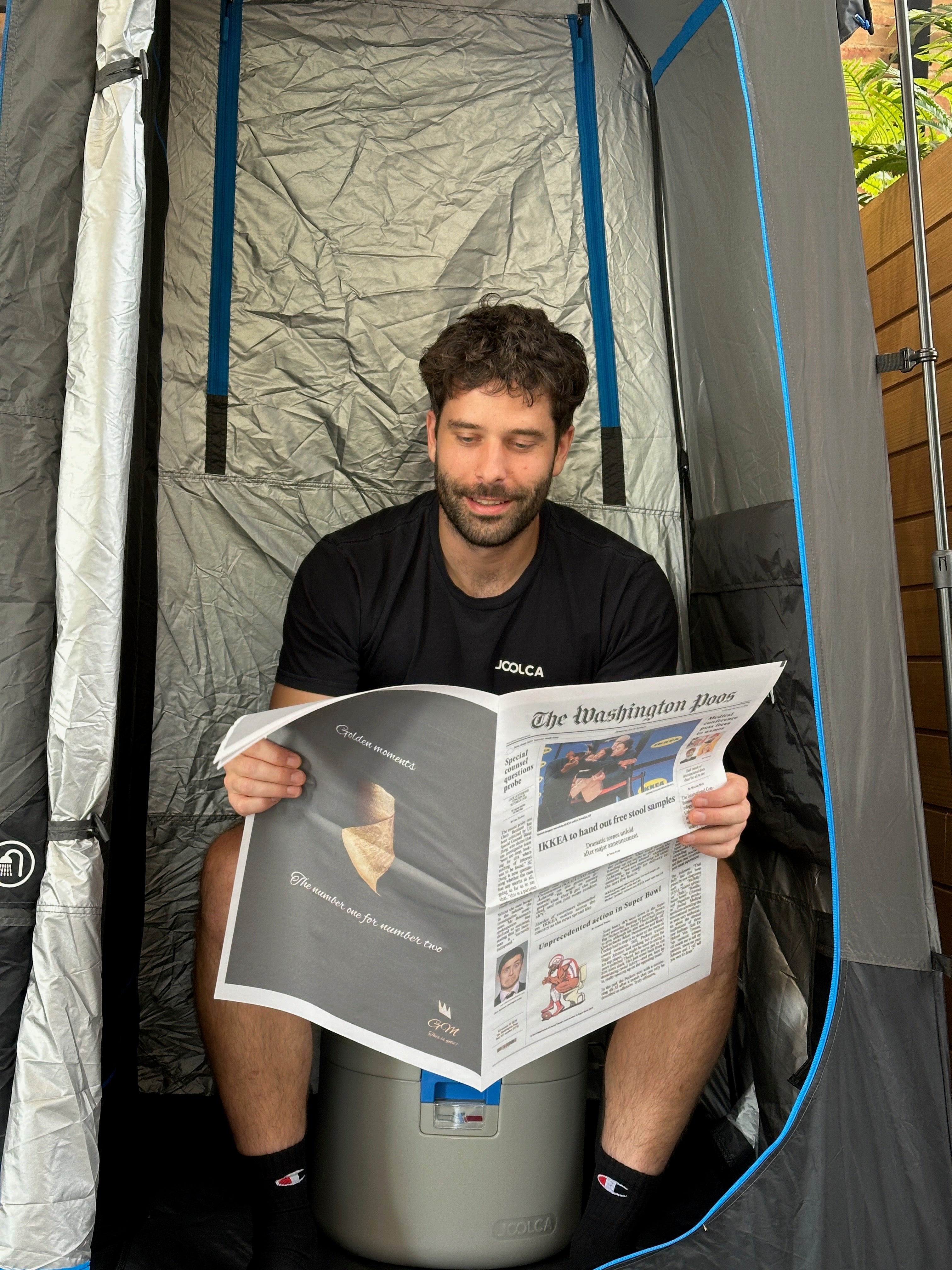 A man sitting inside a portable privacy tent, reading a newspaper while seated on a compact portable toilet, surrounded by outdoor gear.