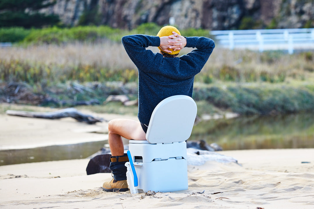 Person in a yellow beanie leaning back on a GottaGo portable toilet on a sandy riverbank, hands behind their head