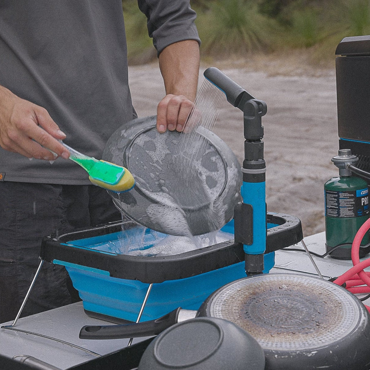 Person using a brush and portable water system to clean a frying pan outdoors, with camping equipment and a portable water heater visible nearby.