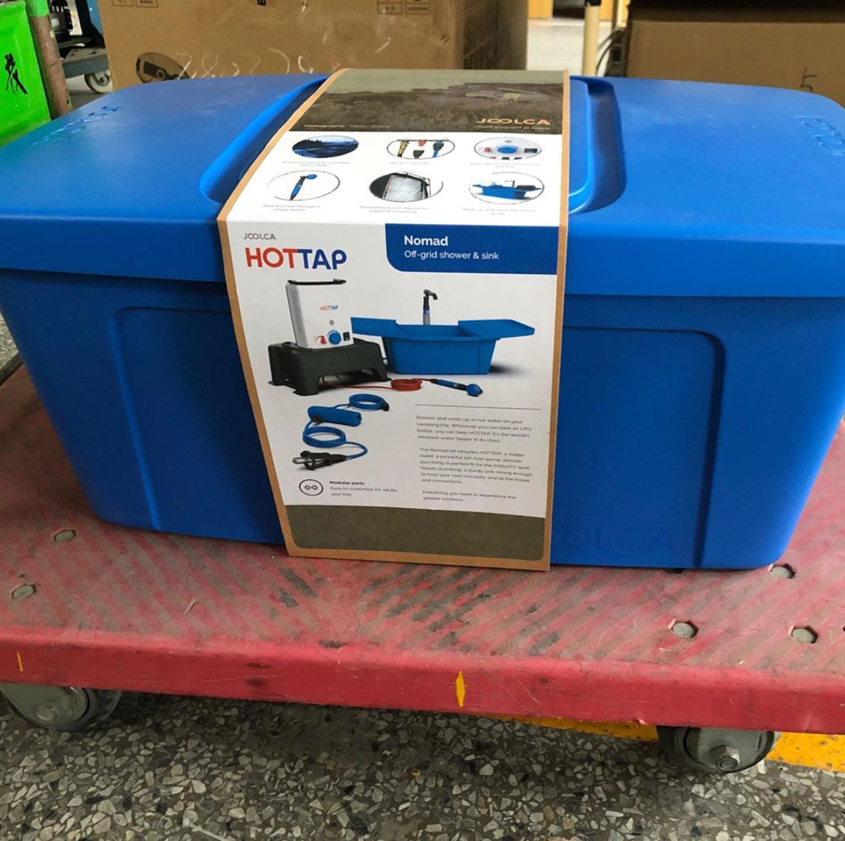 Blue portable container with a label showing components of an off-grid shower and sink system, placed on a red wheeled platform in an industrial setting.