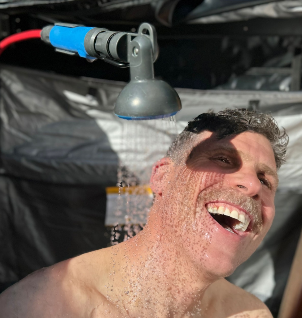 Man taking a shower outdoors with a portable shower head attached to a vehicle.