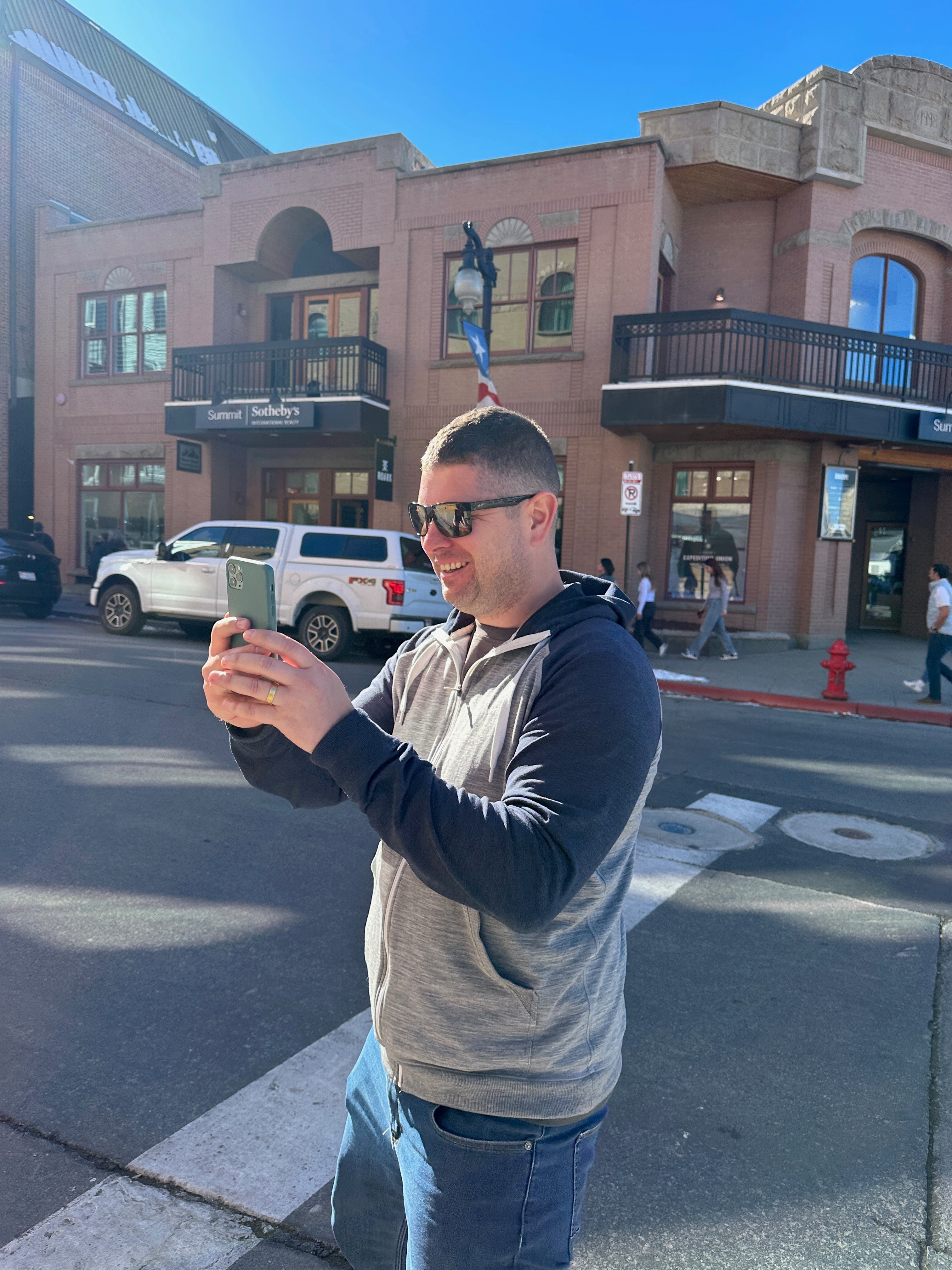 Man wearing sunglasses and a two-tone hoodie standing on a city street in bright sunlight, holding a smartphone and smiling while taking a picture or video.