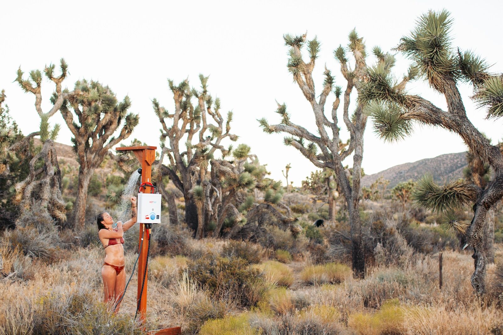 A woman adjusts an outdoor hot shower setup among dry, spiky Joshua trees in a desert landscape, illustrating off-grid portable camping shower solutions.