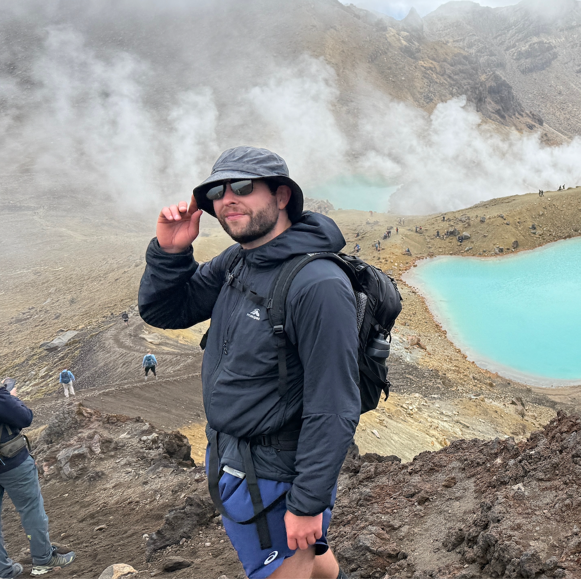 Man wearing outdoor gear and sunglasses standing on rocky terrain near a steaming volcanic crater lake with turquoise water and misty mountain background.