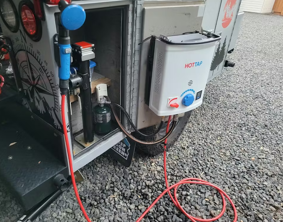 Mounted portable hot water system attached to an outdoor vehicle with hoses connected to a water source and propane tank for off-grid hot water supply.