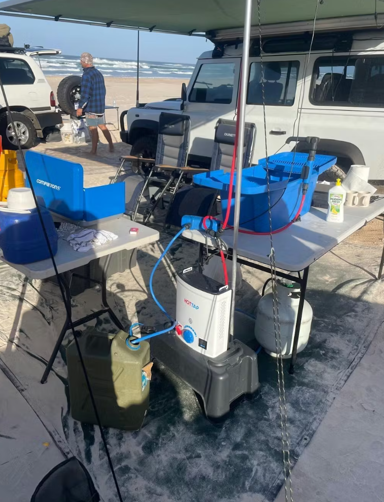 Outdoor beach setup with portable instant hot water system connected to a fuel source, folding tables with camping supplies, chairs, and two off-road vehicles parked nearby.