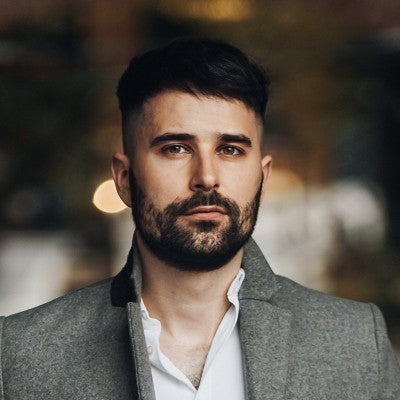 Young man with dark hair and beard wearing a grey coat and white shirt, standing indoors with a softly blurred background.
