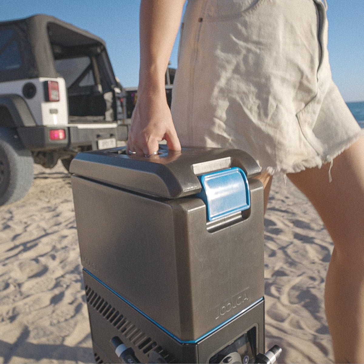 Person carrying a portable outdoor water heater on a sandy beach next to an off-road vehicle, featuring the HOTTAP Go with self-contained tank and pump for instant hot water camping.