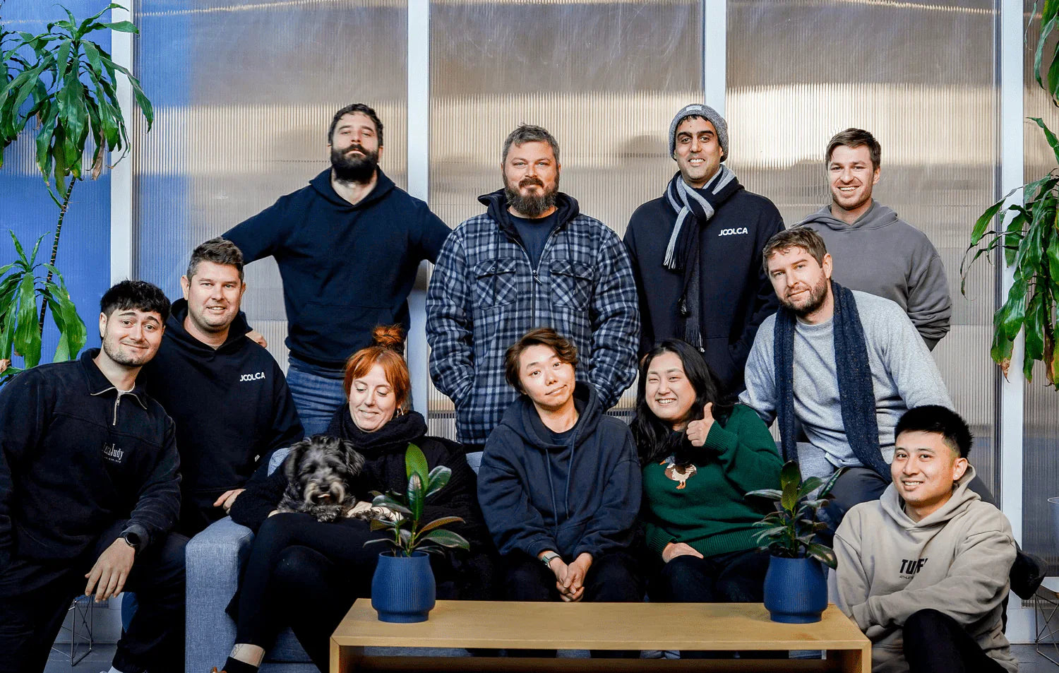 Group of eleven people sitting and standing indoors, casually dressed and smiling, with two potted plants and a small dog on a coffee table in front of them.
