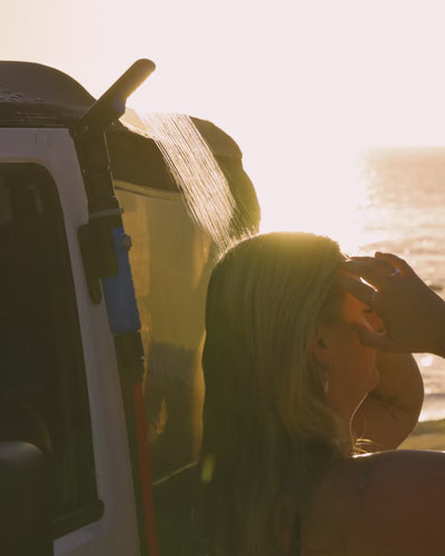 Woman rinsing sand from her hair using a HOTTAP Go outdoor shower at sunset on the beach.